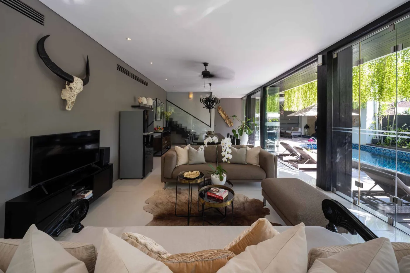 Modern dining room in a tropical villa with a set wooden table, chandelier, and glass walls overlooking a lit infinity pool and garden at night. - Bali Villas