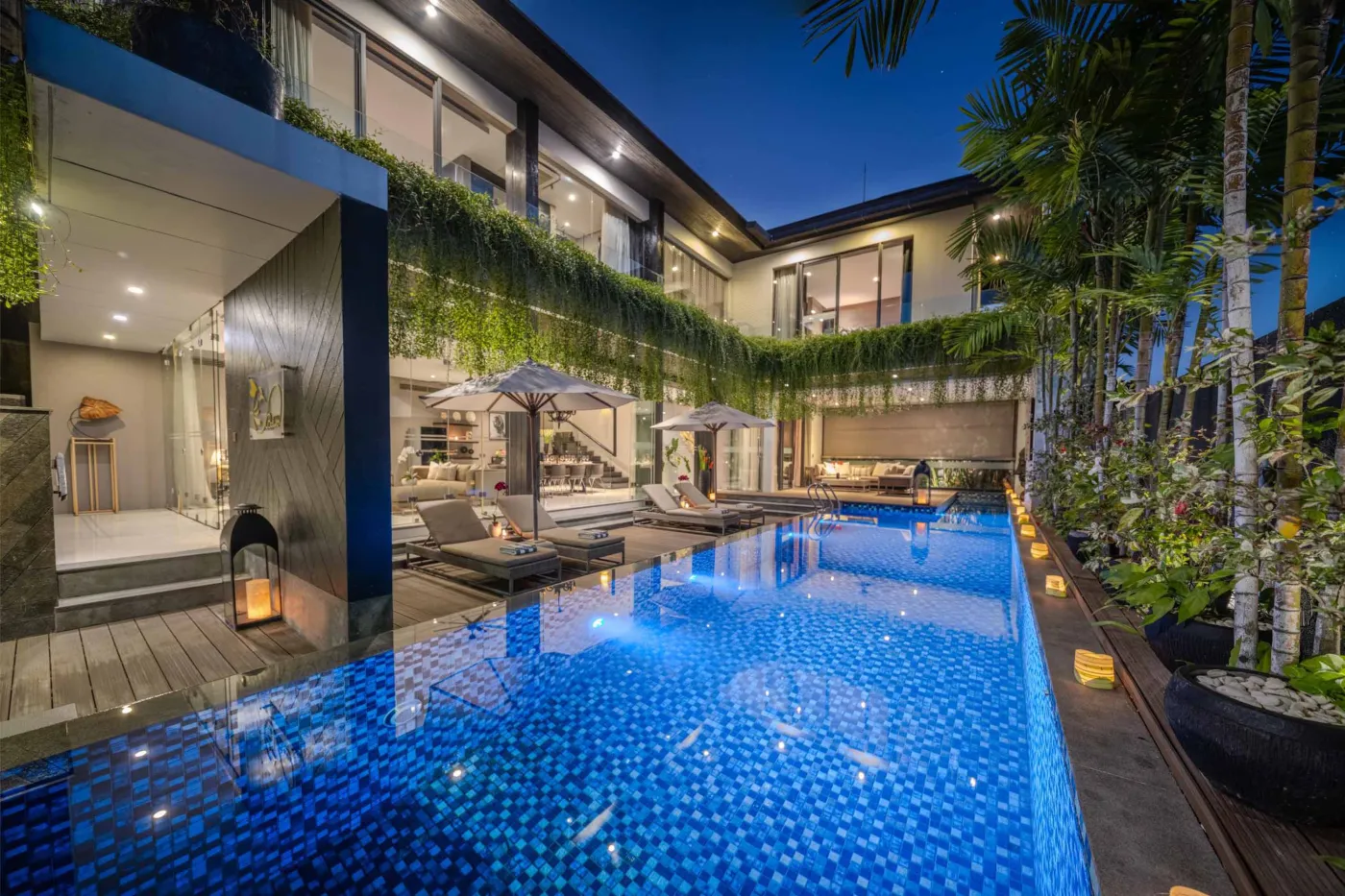 Modern dining room in a tropical villa with a set wooden table, chandelier, and glass walls overlooking a lit infinity pool and garden at night. - Bali Villas