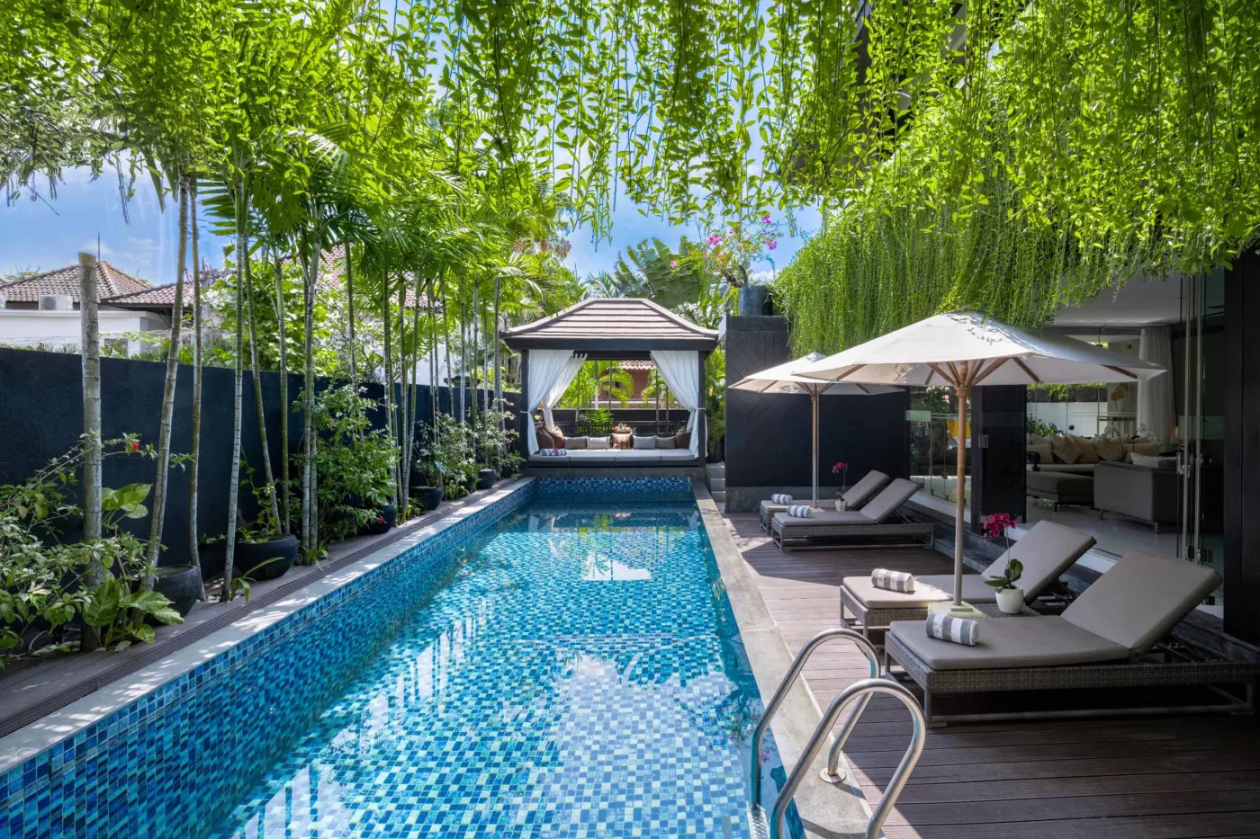 Modern dining room in a tropical villa with a set wooden table, chandelier, and glass walls overlooking a lit infinity pool and garden at night. - Bali Villas