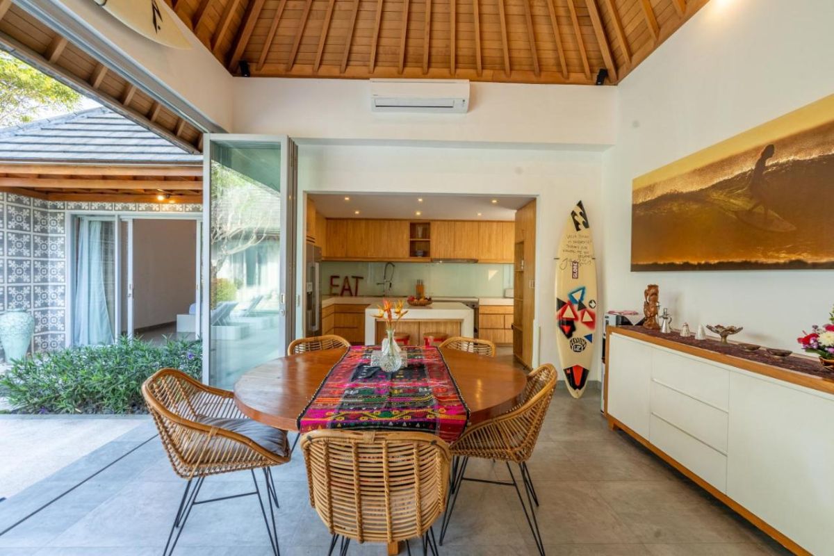Spacious Balinese villa interior with open-plan living-dining area, wooden ceiling, wicker dining table set with colorful food, lounge sofas, and sliding doors to tropical pool garden. - Bali Villas