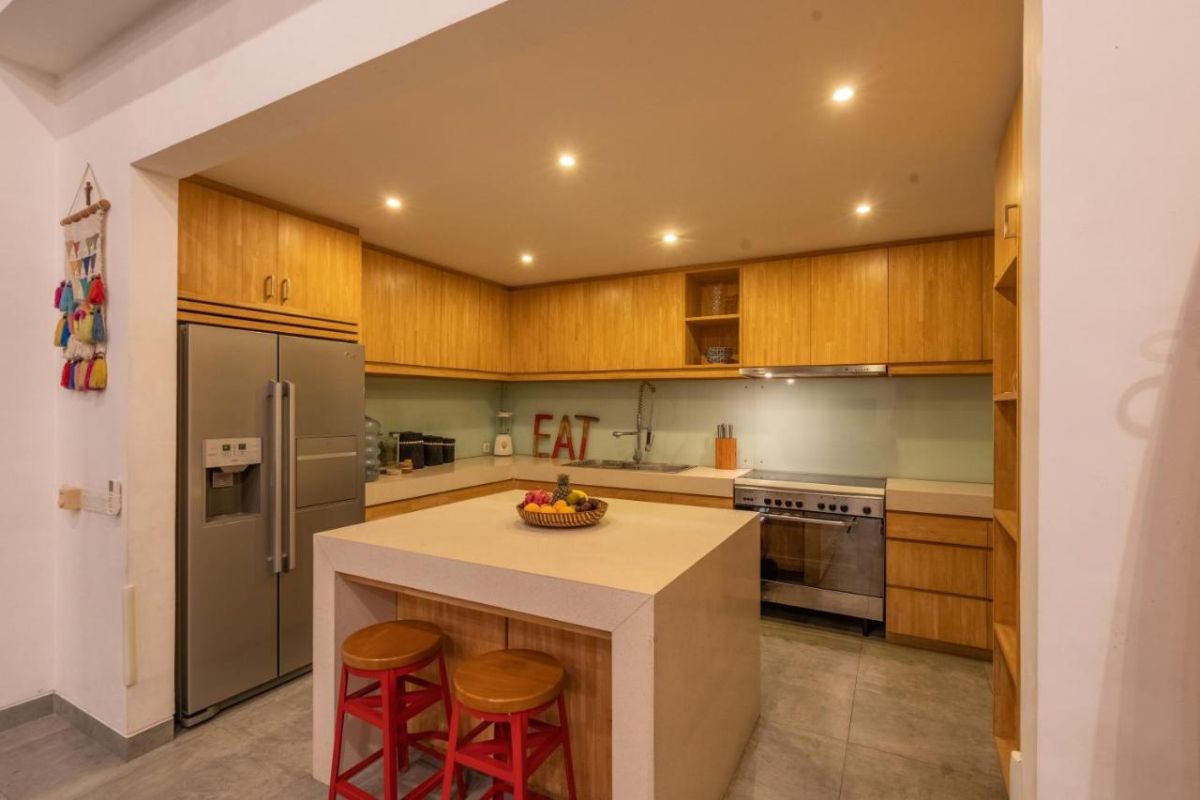 Spacious Balinese villa interior with open-plan living-dining area, wooden ceiling, wicker dining table set with colorful food, lounge sofas, and sliding doors to tropical pool garden. - Bali Villas