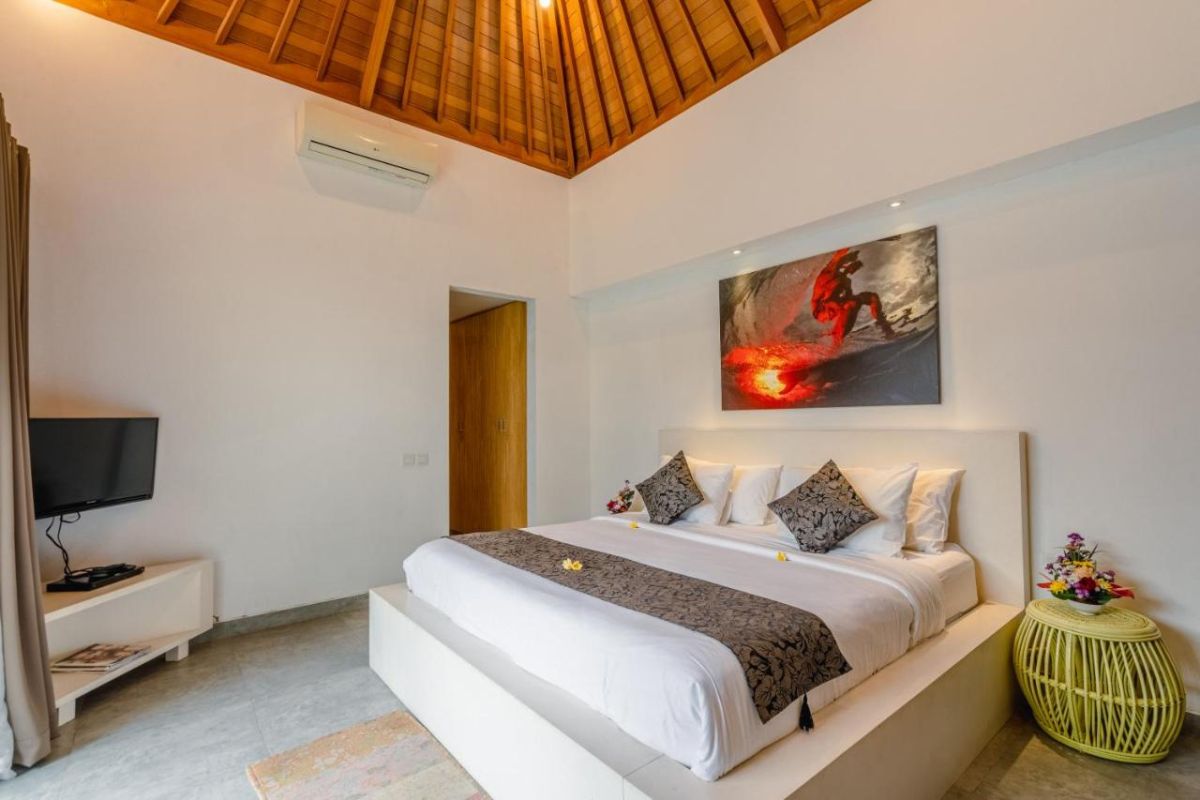 Spacious Balinese villa interior with open-plan living-dining area, wooden ceiling, wicker dining table set with colorful food, lounge sofas, and sliding doors to tropical pool garden. - Bali Villas