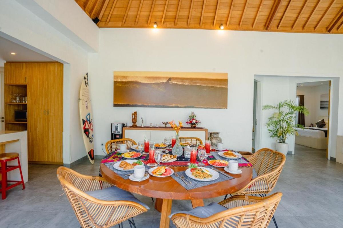 Spacious Balinese villa interior with open-plan living-dining area, wooden ceiling, wicker dining table set with colorful food, lounge sofas, and sliding doors to tropical pool garden. - Bali Villas