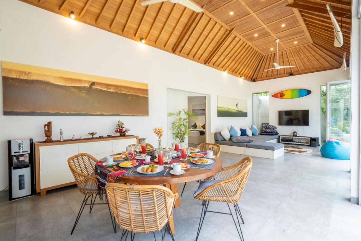 Spacious Balinese villa interior with open-plan living-dining area, wooden ceiling, wicker dining table set with colorful food, lounge sofas, and sliding doors to tropical pool garden. - Bali Villas