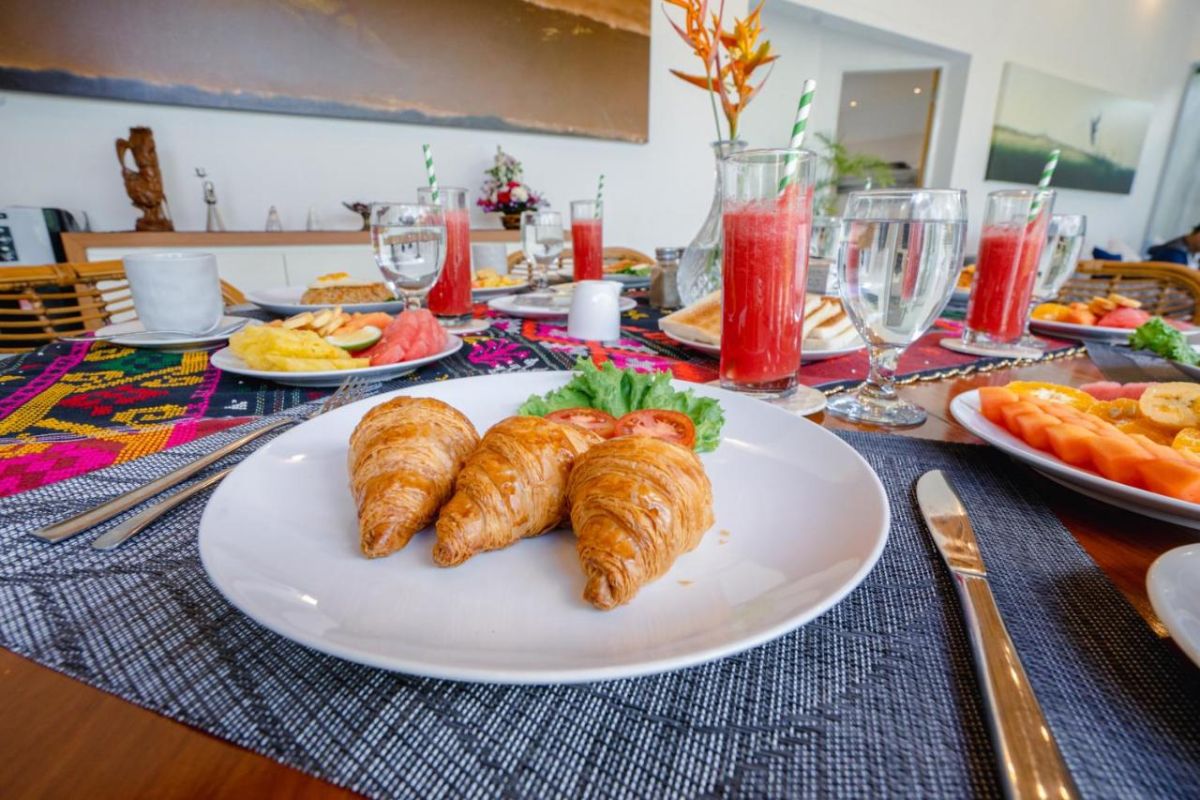 Spacious Balinese villa interior with open-plan living-dining area, wooden ceiling, wicker dining table set with colorful food, lounge sofas, and sliding doors to tropical pool garden. - Bali Villas