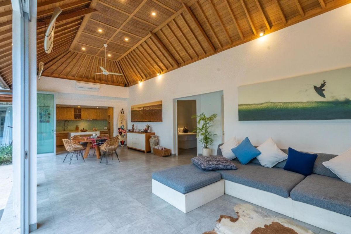 Spacious Balinese villa interior with open-plan living-dining area, wooden ceiling, wicker dining table set with colorful food, lounge sofas, and sliding doors to tropical pool garden. - Bali Villas