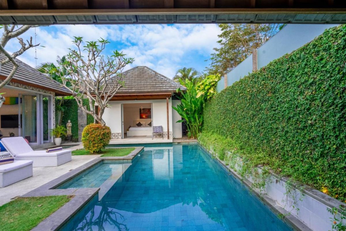 Spacious Balinese villa interior with open-plan living-dining area, wooden ceiling, wicker dining table set with colorful food, lounge sofas, and sliding doors to tropical pool garden. - Bali Villas