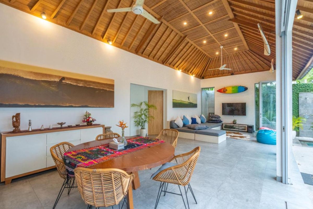 Spacious Balinese villa interior with open-plan living-dining area, wooden ceiling, wicker dining table set with colorful food, lounge sofas, and sliding doors to tropical pool garden. - Bali Villas