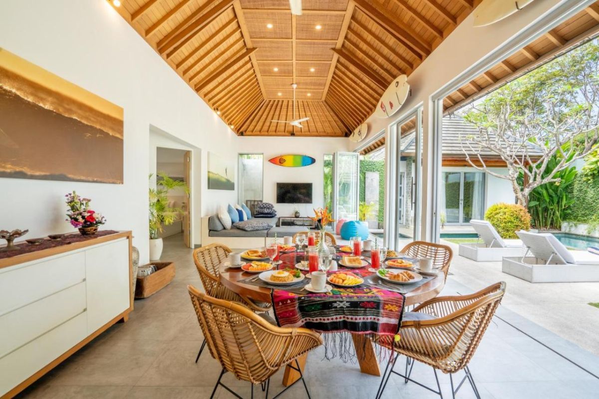 Spacious Balinese villa interior with open-plan living-dining area, wooden ceiling, wicker dining table set with colorful food, lounge sofas, and sliding doors to tropical pool garden. - Bali Villas
