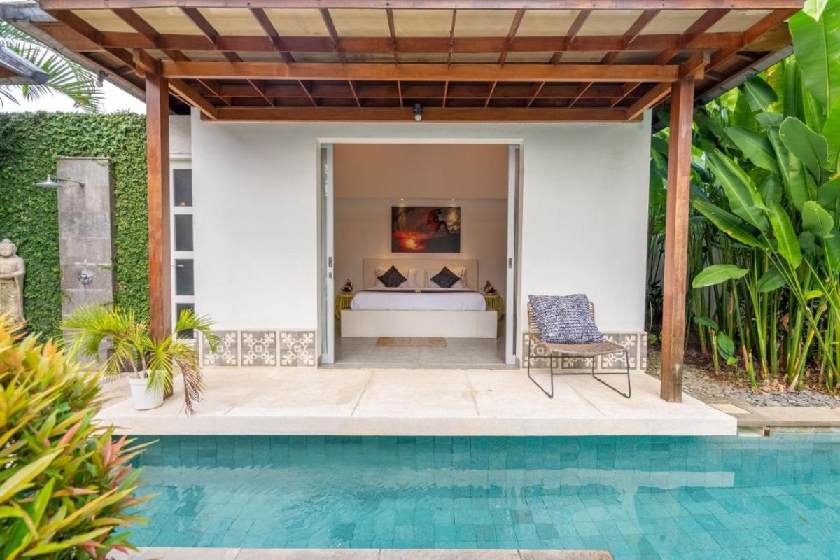 Spacious Balinese villa interior with open-plan living-dining area, wooden ceiling, wicker dining table set with colorful food, lounge sofas, and sliding doors to tropical pool garden. - Bali Villas