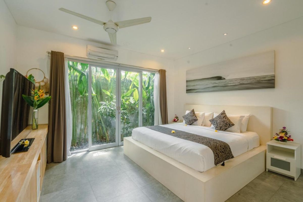Spacious Balinese villa interior with open-plan living-dining area, wooden ceiling, wicker dining table set with colorful food, lounge sofas, and sliding doors to tropical pool garden. - Bali Villas