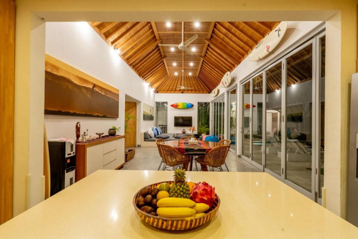 Spacious Balinese villa interior with open-plan living-dining area, wooden ceiling, wicker dining table set with colorful food, lounge sofas, and sliding doors to tropical pool garden. - Bali Villas