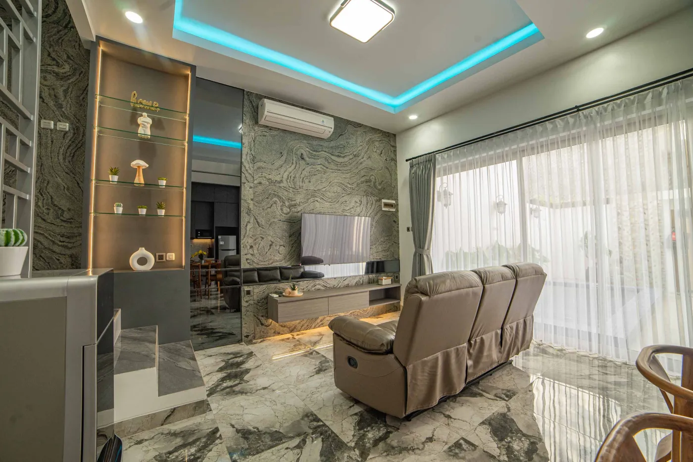 Modern living room interior with beige L-shaped sofa, wall-mounted TV, blue LED ceiling lights, textured gray wall, and potted plants. - Bali Villas