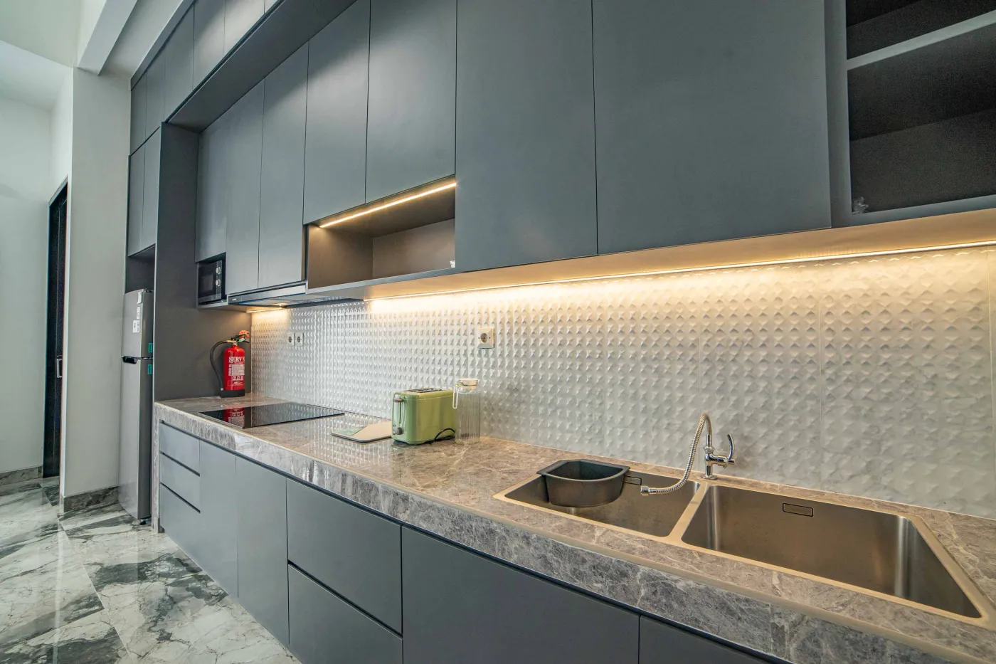 Modern living room interior with beige L-shaped sofa, wall-mounted TV, blue LED ceiling lights, textured gray wall, and potted plants. - Bali Villas