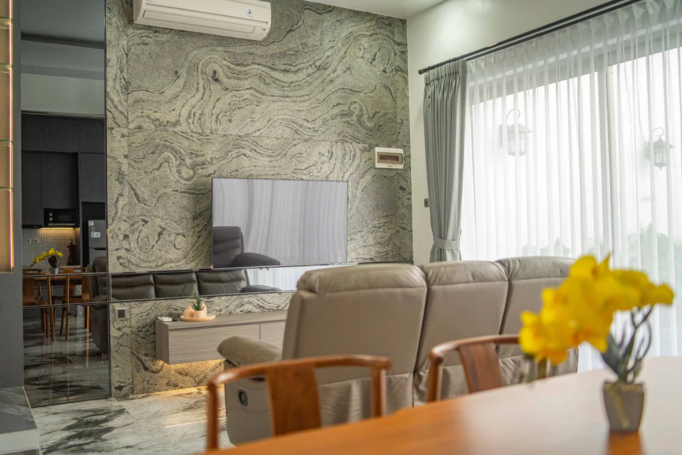 Modern living room interior with beige L-shaped sofa, wall-mounted TV, blue LED ceiling lights, textured gray wall, and potted plants. - Bali Villas