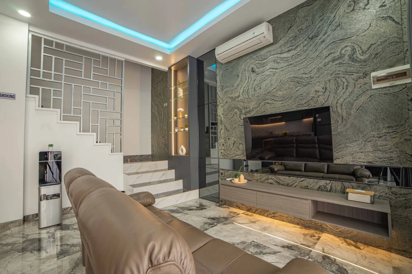 Modern living room interior with beige L-shaped sofa, wall-mounted TV, blue LED ceiling lights, textured gray wall, and potted plants. - Bali Villas