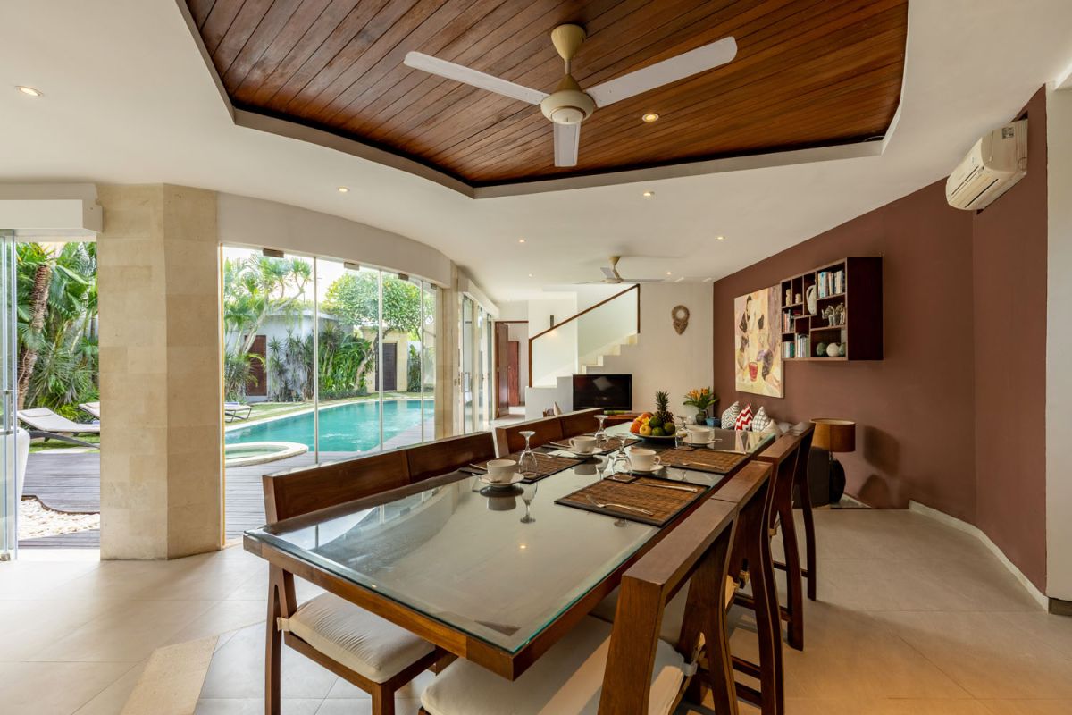 Modern Balinese kitchen interior with white cabinets, marble island, wooden bar stools, pendant lights, and tropical greenery. - Bali Villas