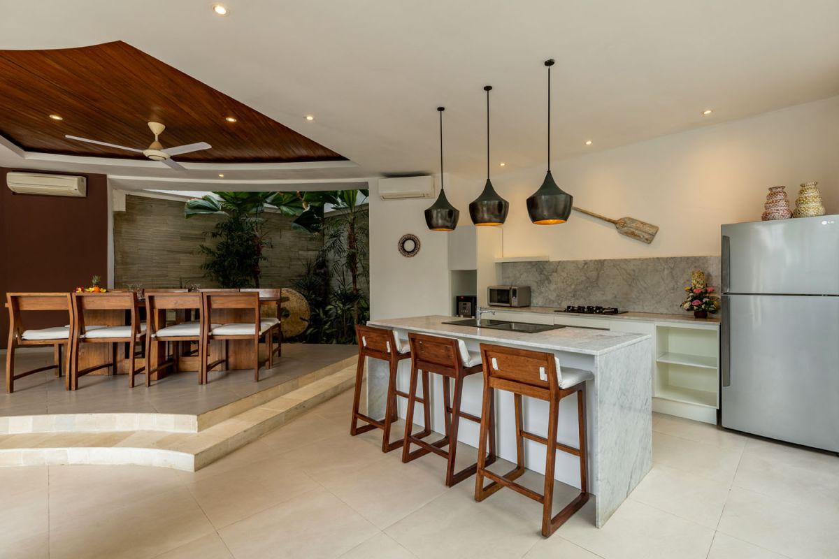 Modern Balinese kitchen interior with white cabinets, marble island, wooden bar stools, pendant lights, and tropical greenery. - Bali Villas