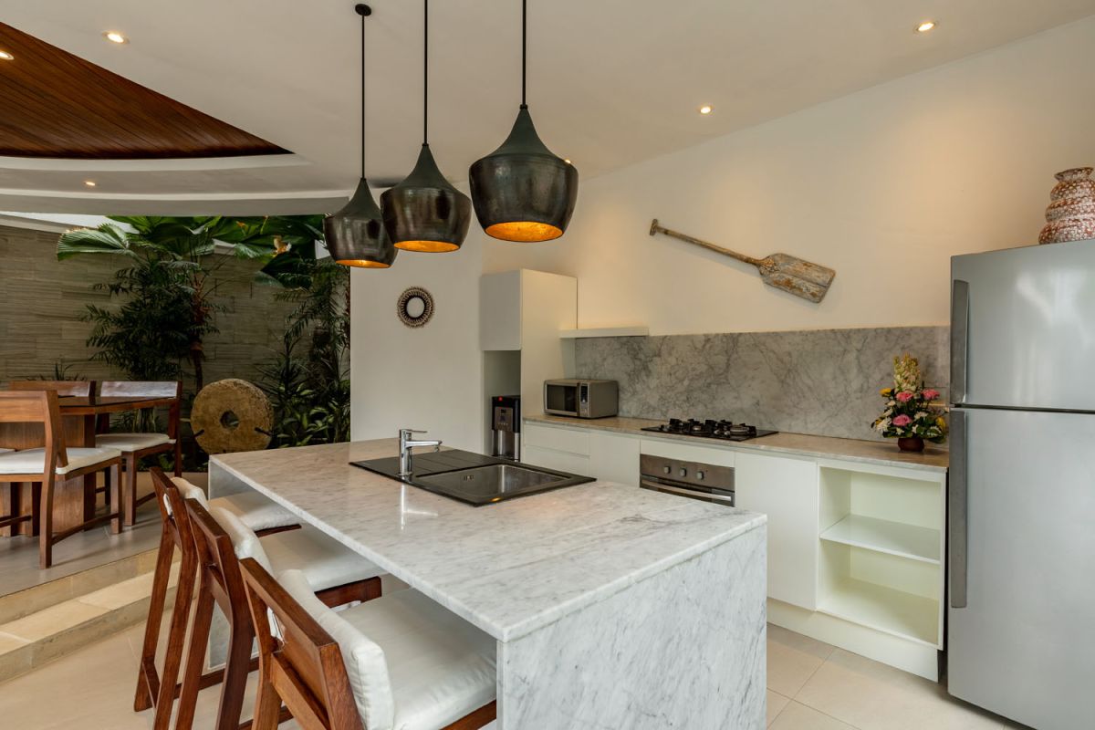 Modern Balinese kitchen interior with white cabinets, marble island, wooden bar stools, pendant lights, and tropical greenery. - Bali Villas