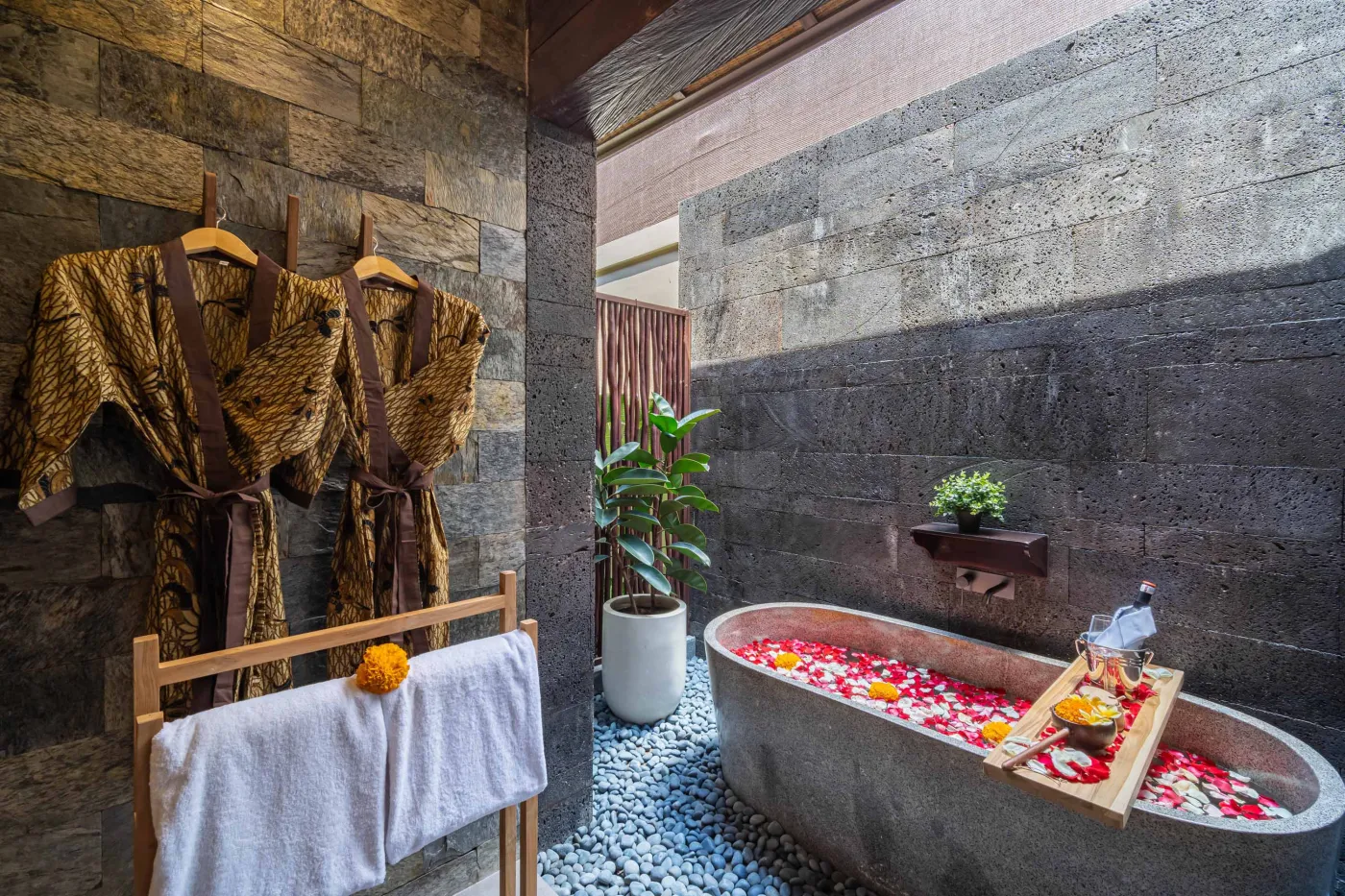 Luxurious outdoor Balinese bathtub with rose petals, tropical flowers, and champagne, surrounded by stone walls, hanging batik robes, and potted plants. - Bali Villas