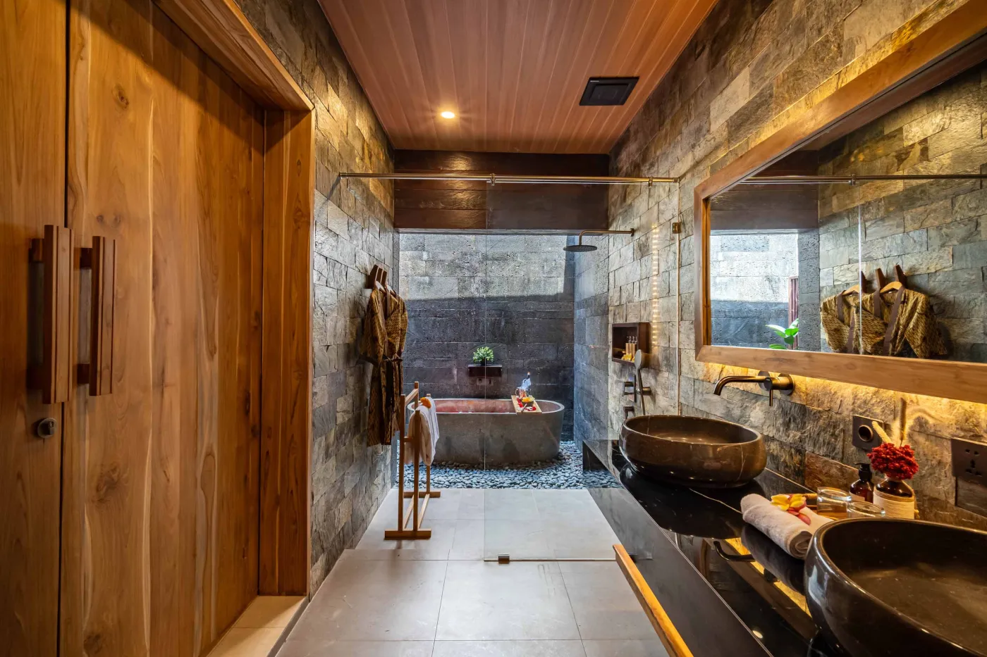 Luxurious outdoor Balinese bathtub with rose petals, tropical flowers, and champagne, surrounded by stone walls, hanging batik robes, and potted plants. - Bali Villas