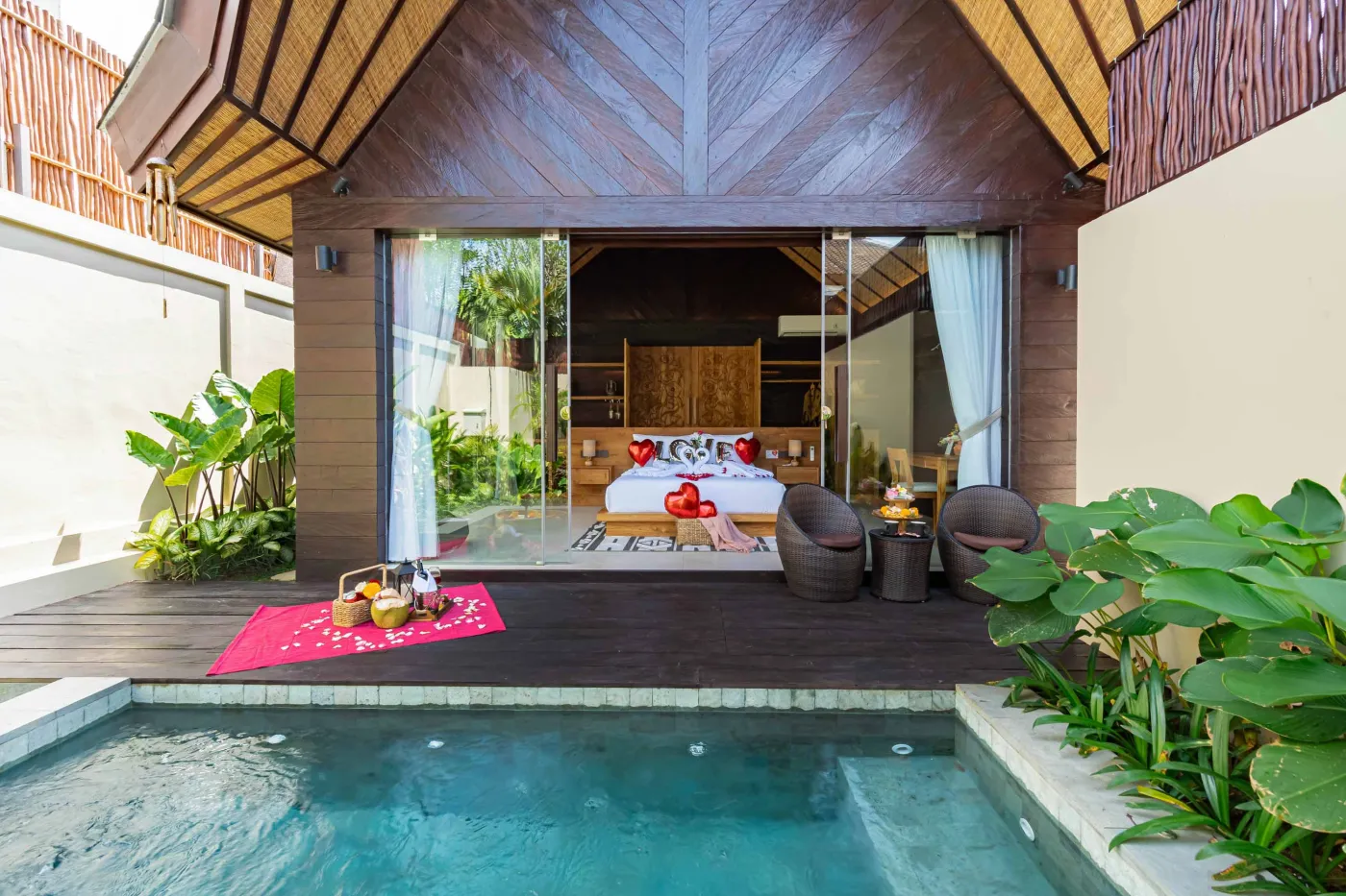 Luxurious outdoor Balinese bathtub with rose petals, tropical flowers, and champagne, surrounded by stone walls, hanging batik robes, and potted plants. - Bali Villas