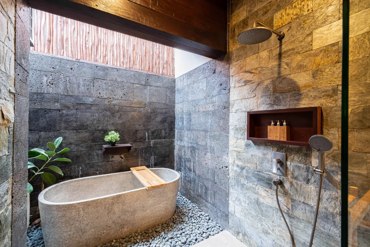 Luxurious outdoor Balinese bathtub with rose petals, tropical flowers, and champagne, surrounded by stone walls, hanging batik robes, and potted plants. - Bali Villas