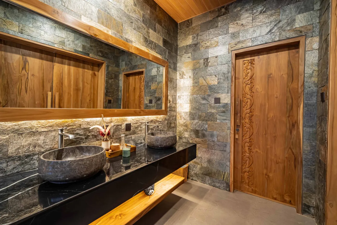 Luxurious outdoor Balinese bathtub with rose petals, tropical flowers, and champagne, surrounded by stone walls, hanging batik robes, and potted plants. - Bali Villas