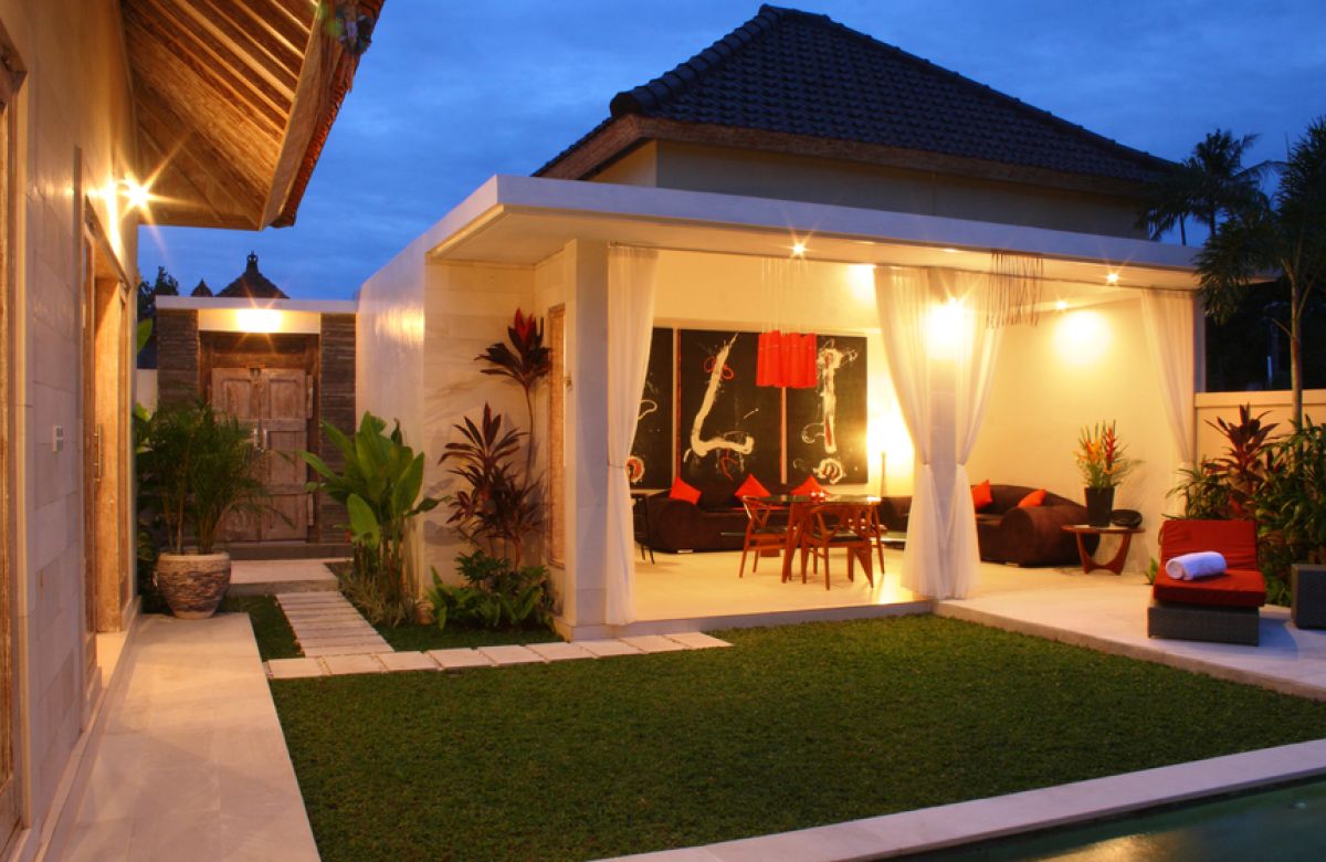 Luxurious Balinese villa interior at night: open dining area with glass table, wooden chairs, red lounger, pendant lamps, and garden pool view through white curtains. - Bali Villas