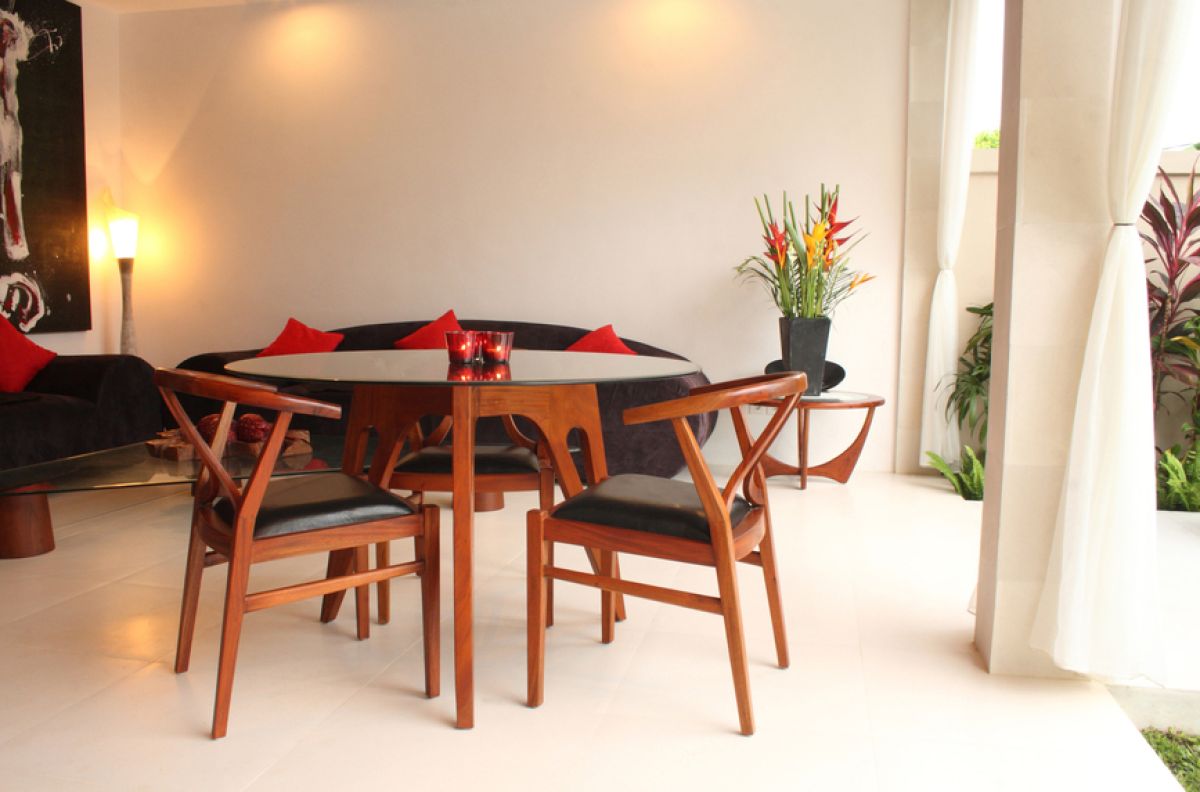 Luxurious Balinese villa interior at night: open dining area with glass table, wooden chairs, red lounger, pendant lamps, and garden pool view through white curtains. - Bali Villas