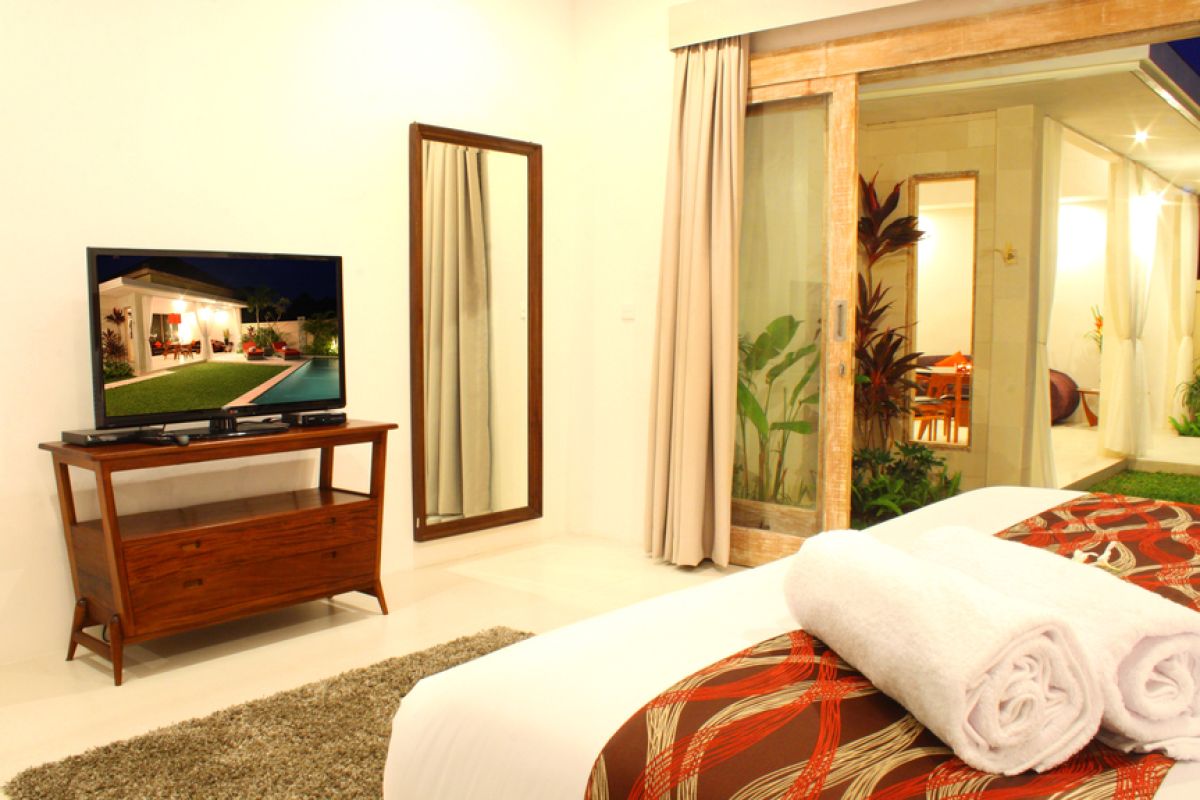 Luxurious Balinese villa interior at night: open dining area with glass table, wooden chairs, red lounger, pendant lamps, and garden pool view through white curtains. - Bali Villas