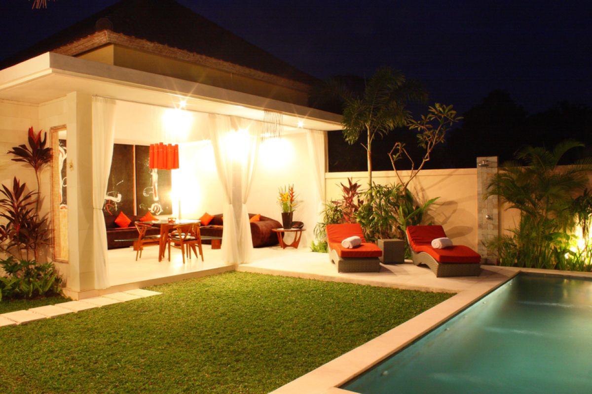 Luxurious Balinese villa interior at night: open dining area with glass table, wooden chairs, red lounger, pendant lamps, and garden pool view through white curtains. - Bali Villas