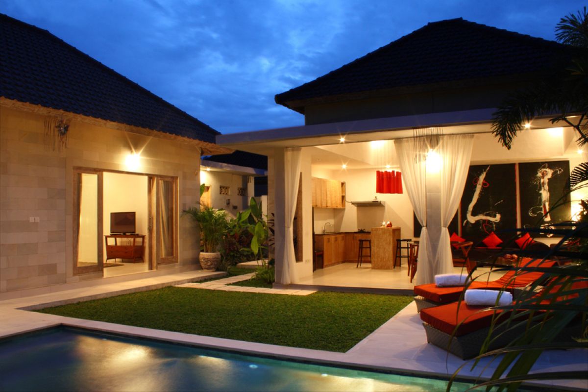 Luxurious Balinese villa interior at night: open dining area with glass table, wooden chairs, red lounger, pendant lamps, and garden pool view through white curtains. - Bali Villas