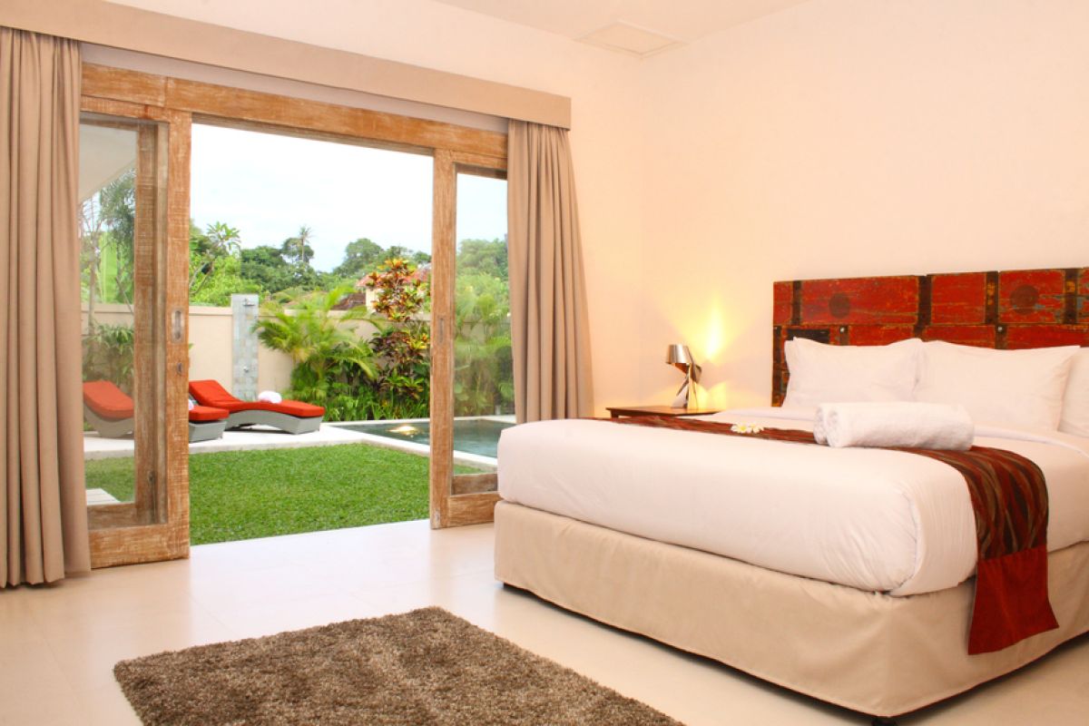 Luxurious Balinese villa interior at night: open dining area with glass table, wooden chairs, red lounger, pendant lamps, and garden pool view through white curtains. - Bali Villas