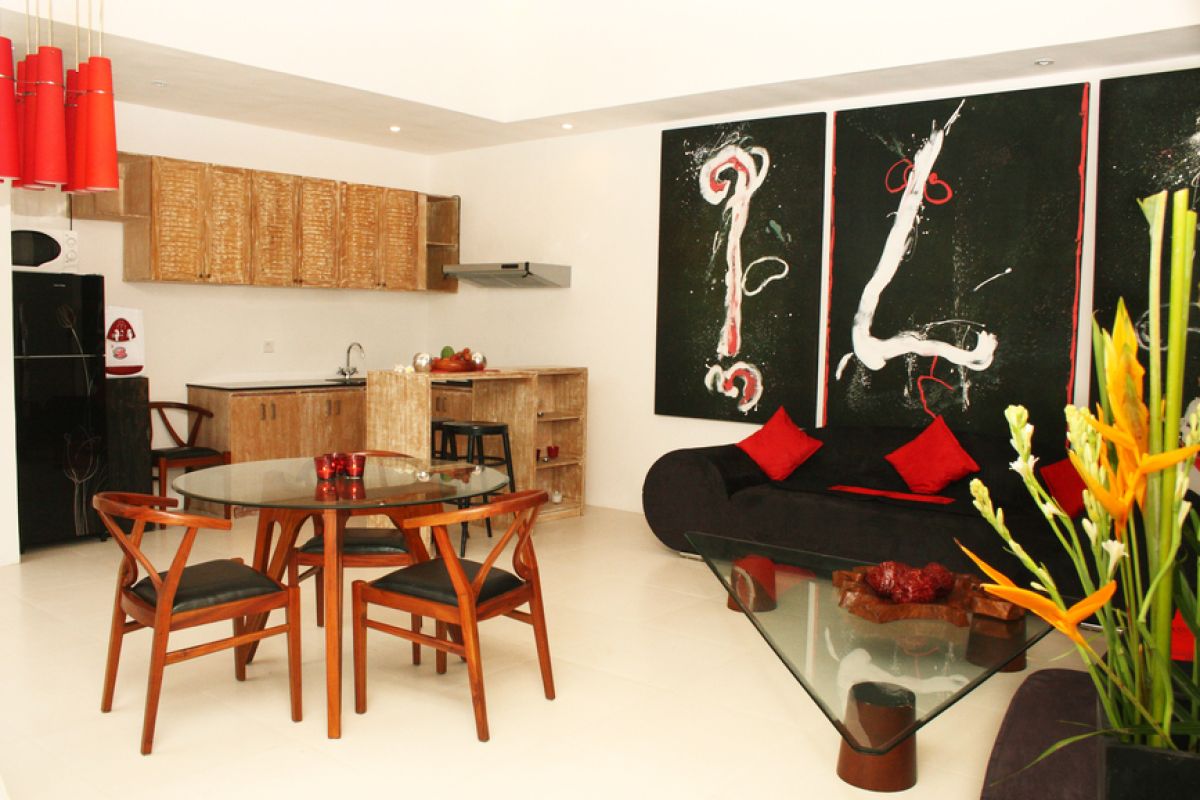 Luxurious Balinese villa interior at night: open dining area with glass table, wooden chairs, red lounger, pendant lamps, and garden pool view through white curtains. - Bali Villas