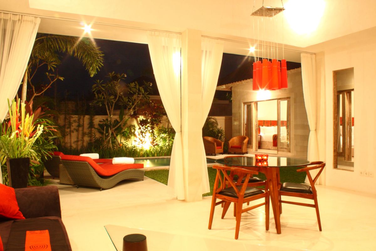 Luxurious Balinese villa interior at night: open dining area with glass table, wooden chairs, red lounger, pendant lamps, and garden pool view through white curtains. - Bali Villas