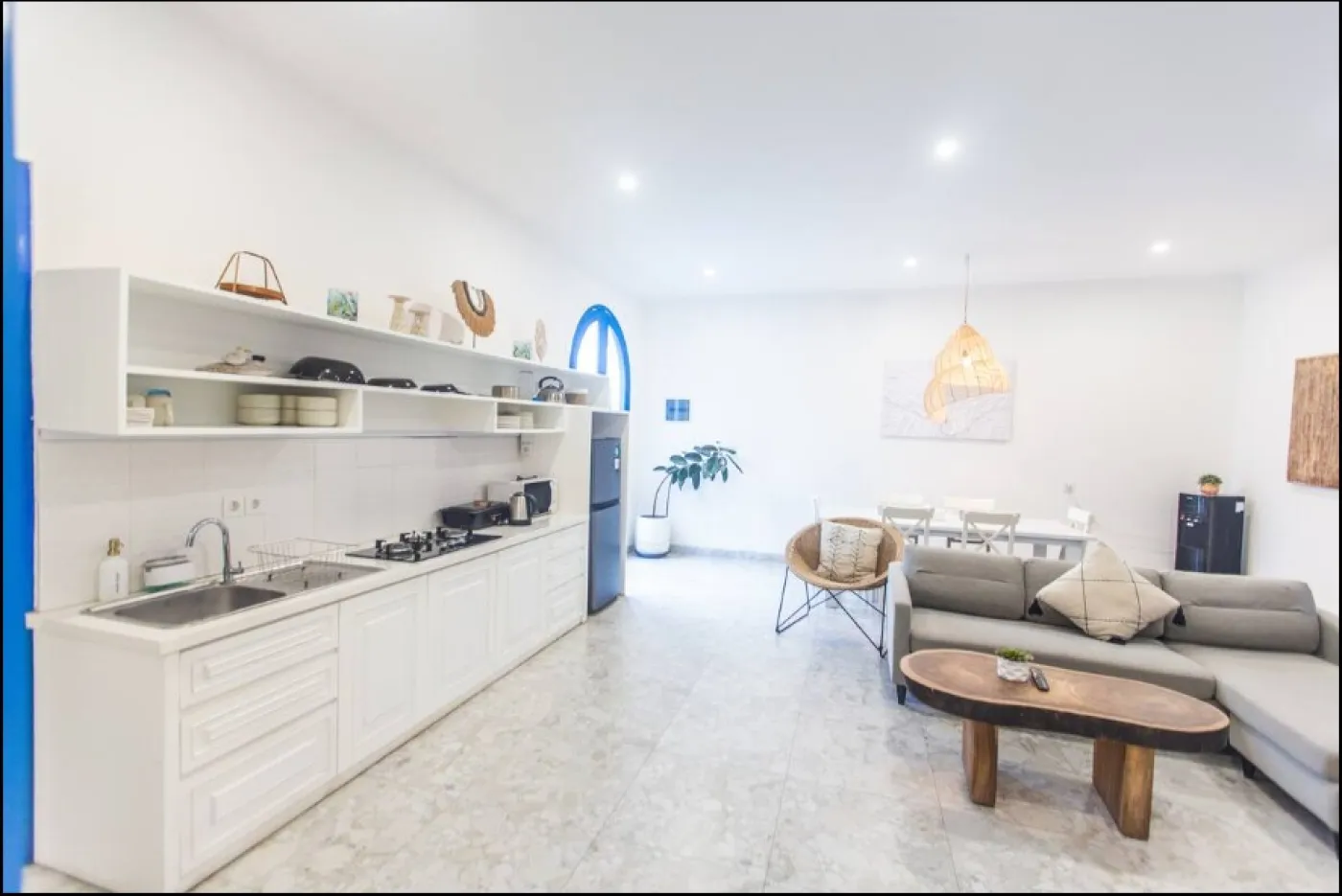 Modern open-plan tropical villa interior with white kitchen cabinets, gray sofa lounge, wooden accents, and potted plants. - Bali Villas