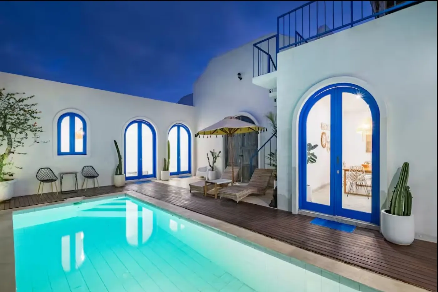 Nighttime view of a luxurious poolside courtyard in a white Mediterranean-style villa with blue arched doors, lounge chairs, potted cacti, and a lit turquoise pool under a starry sky. - Bali Villas