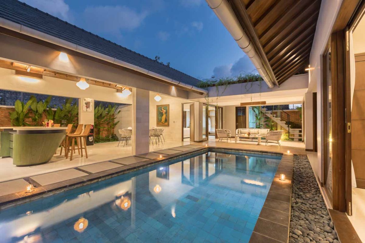 Elegant open-plan dining room in a luxury Balinese villa, featuring a glass-top table with rattan chairs, tropical centerpiece, sleek kitchen, and poolside views. - Bali Villas