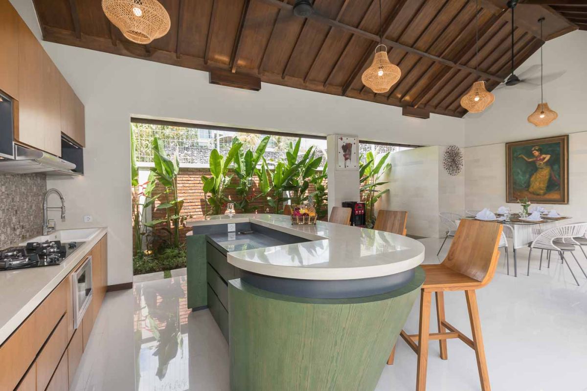 Elegant open-plan dining room in a luxury Balinese villa, featuring a glass-top table with rattan chairs, tropical centerpiece, sleek kitchen, and poolside views. - Bali Villas