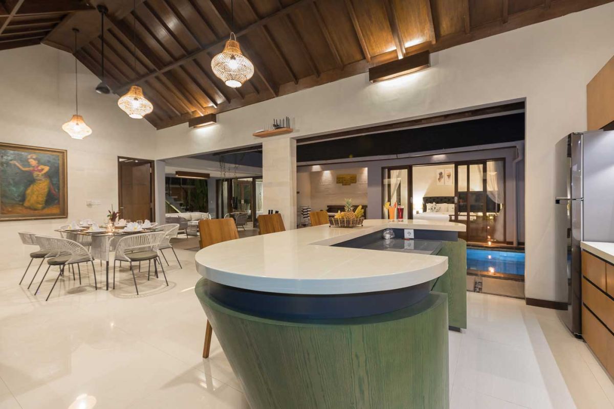 Elegant open-plan dining room in a luxury Balinese villa, featuring a glass-top table with rattan chairs, tropical centerpiece, sleek kitchen, and poolside views. - Bali Villas