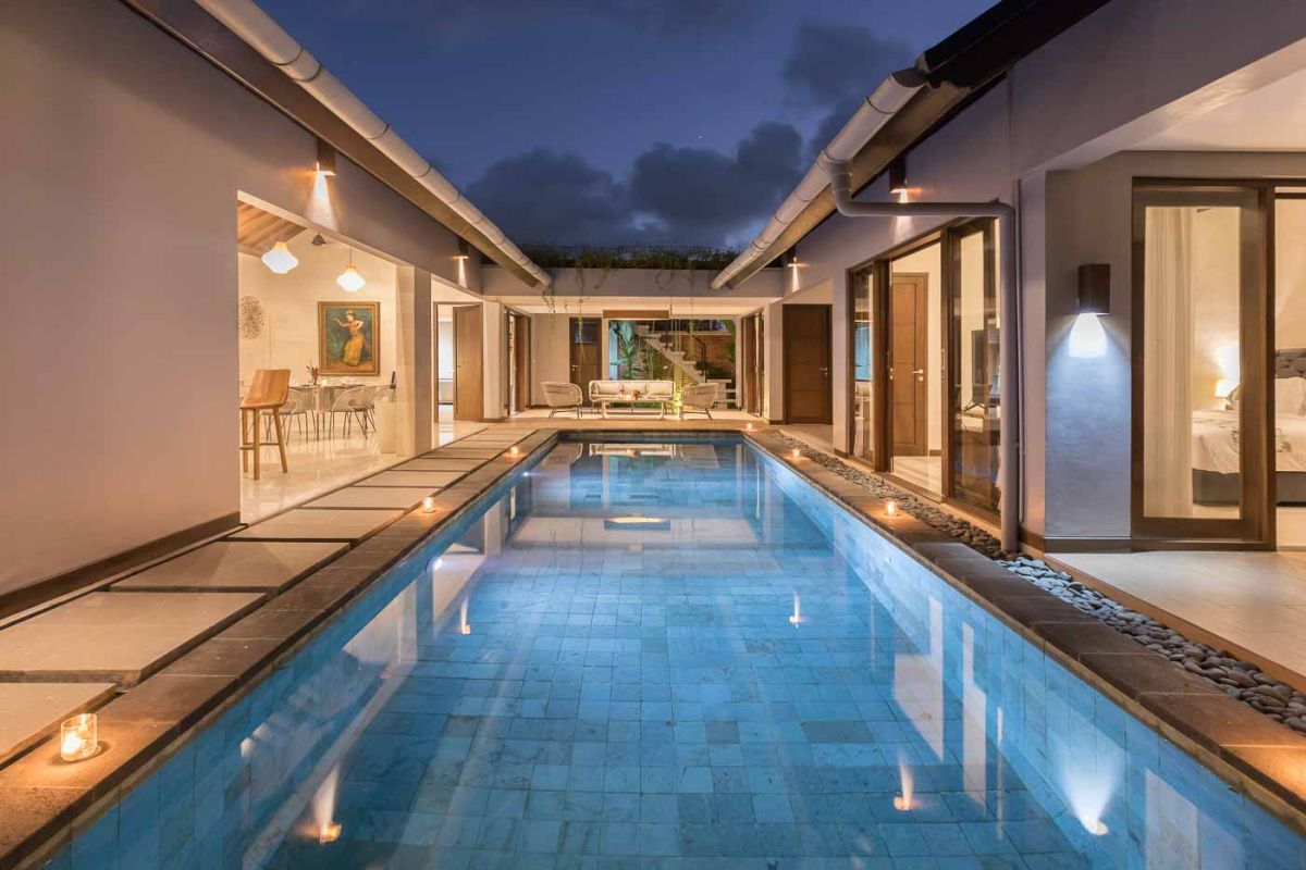 Elegant open-plan dining room in a luxury Balinese villa, featuring a glass-top table with rattan chairs, tropical centerpiece, sleek kitchen, and poolside views. - Bali Villas