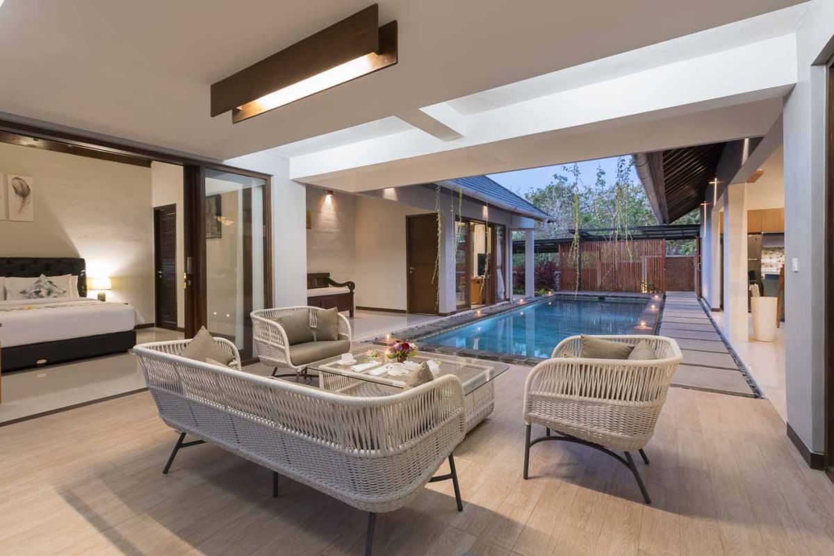 Elegant open-plan dining room in a luxury Balinese villa, featuring a glass-top table with rattan chairs, tropical centerpiece, sleek kitchen, and poolside views. - Bali Villas
