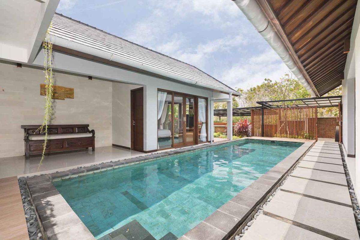 Elegant open-plan dining room in a luxury Balinese villa, featuring a glass-top table with rattan chairs, tropical centerpiece, sleek kitchen, and poolside views. - Bali Villas