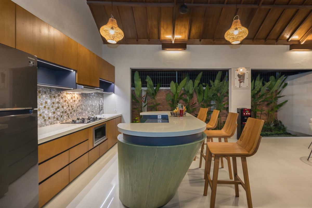 Elegant open-plan dining room in a luxury Balinese villa, featuring a glass-top table with rattan chairs, tropical centerpiece, sleek kitchen, and poolside views. - Bali Villas