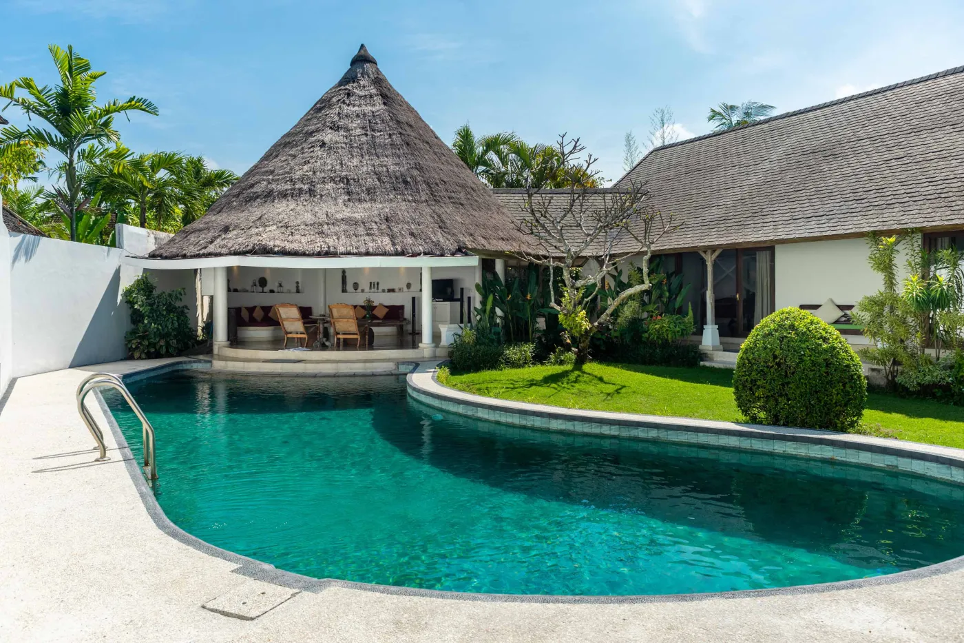 Tropical Bali-style garden with lush palms, stone pathway through green lawn, lounge chairs, blue umbrella, and circular pool beside white wall. - Bali Villas