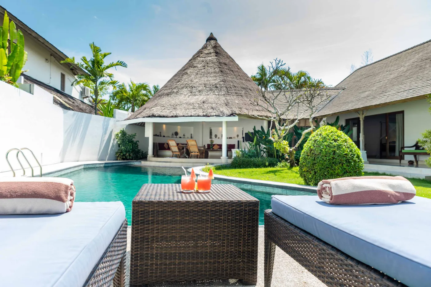 Tropical Bali-style garden with lush palms, stone pathway through green lawn, lounge chairs, blue umbrella, and circular pool beside white wall. - Bali Villas