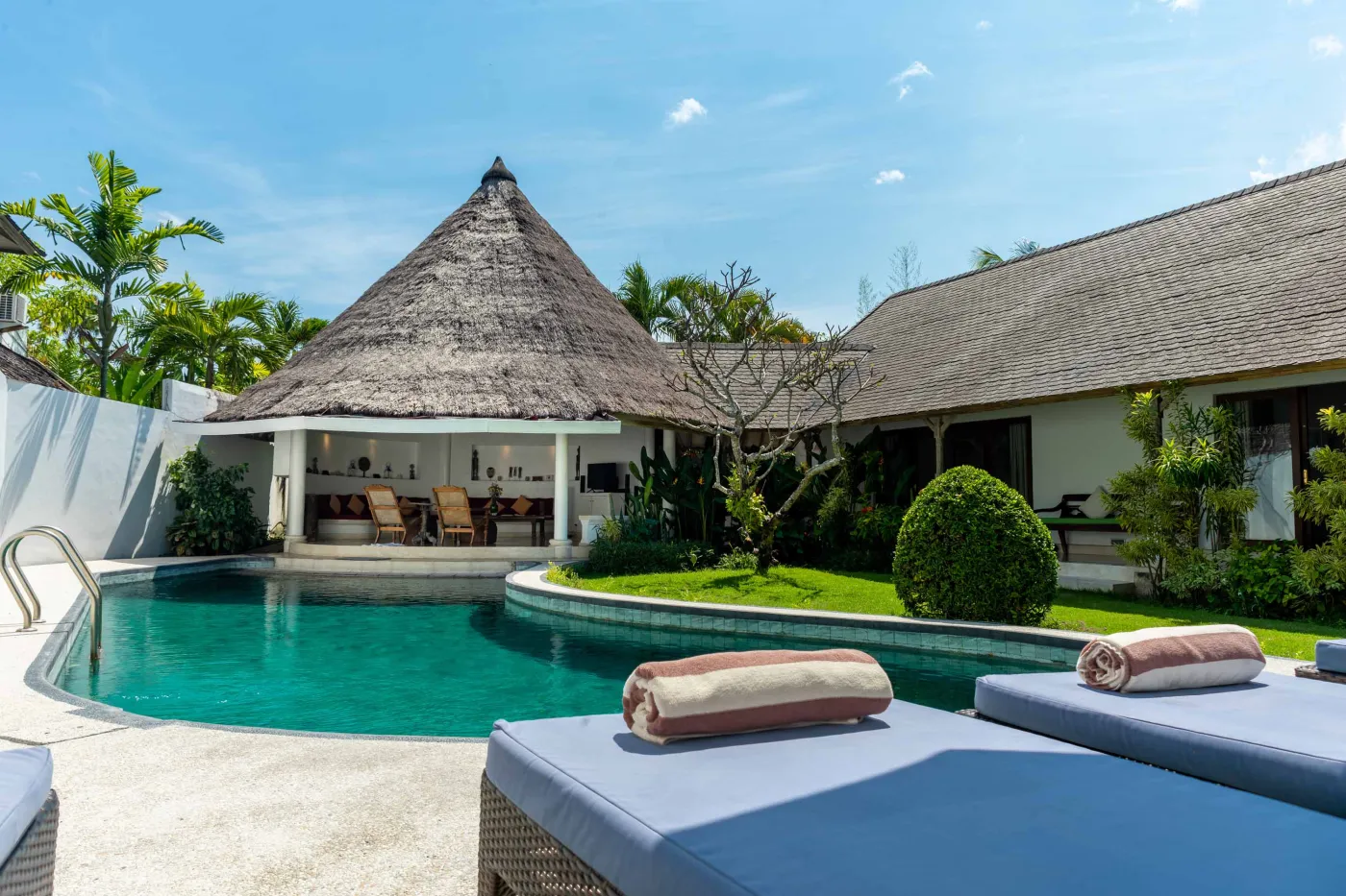 Tropical Bali-style garden with lush palms, stone pathway through green lawn, lounge chairs, blue umbrella, and circular pool beside white wall. - Bali Villas