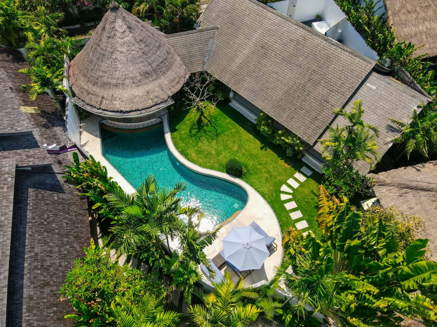 Tropical Bali-style garden with lush palms, stone pathway through green lawn, lounge chairs, blue umbrella, and circular pool beside white wall. - Bali Villas