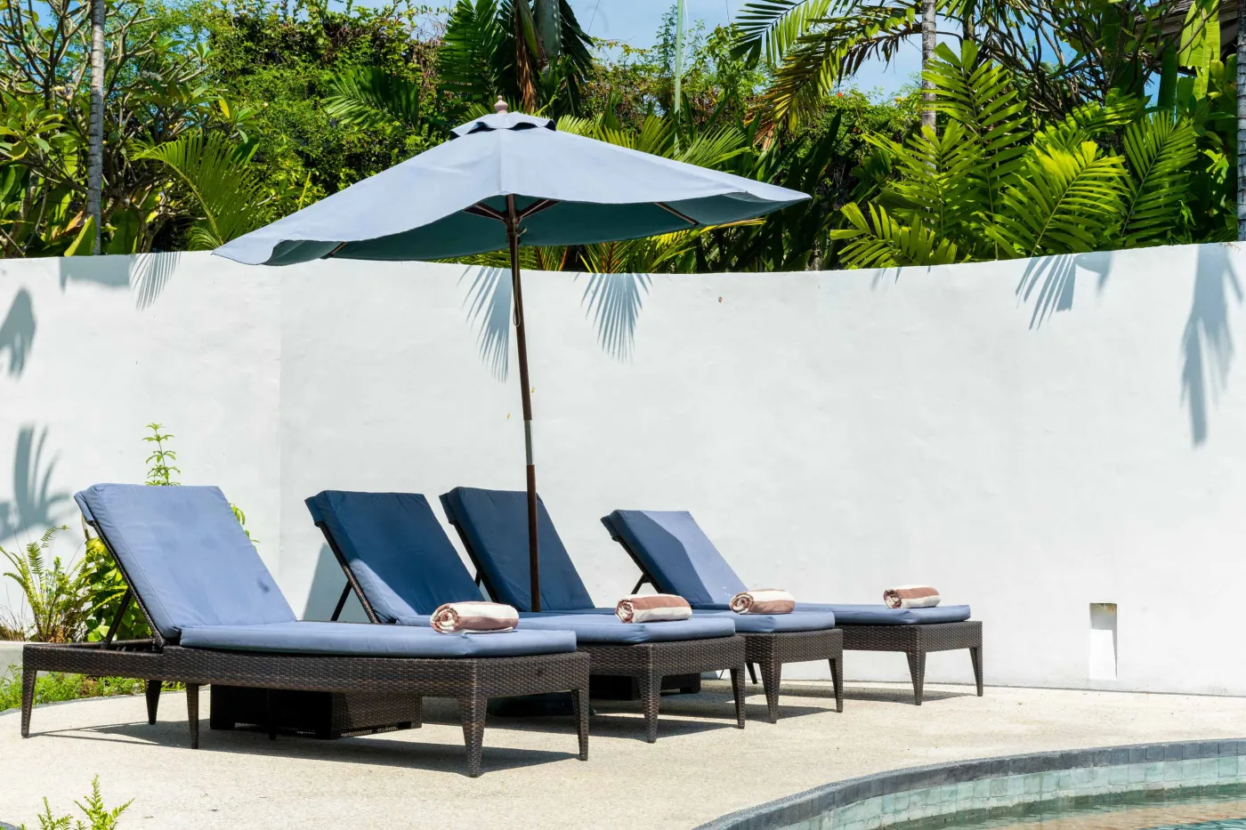 Tropical Bali-style garden with lush palms, stone pathway through green lawn, lounge chairs, blue umbrella, and circular pool beside white wall. - Bali Villas